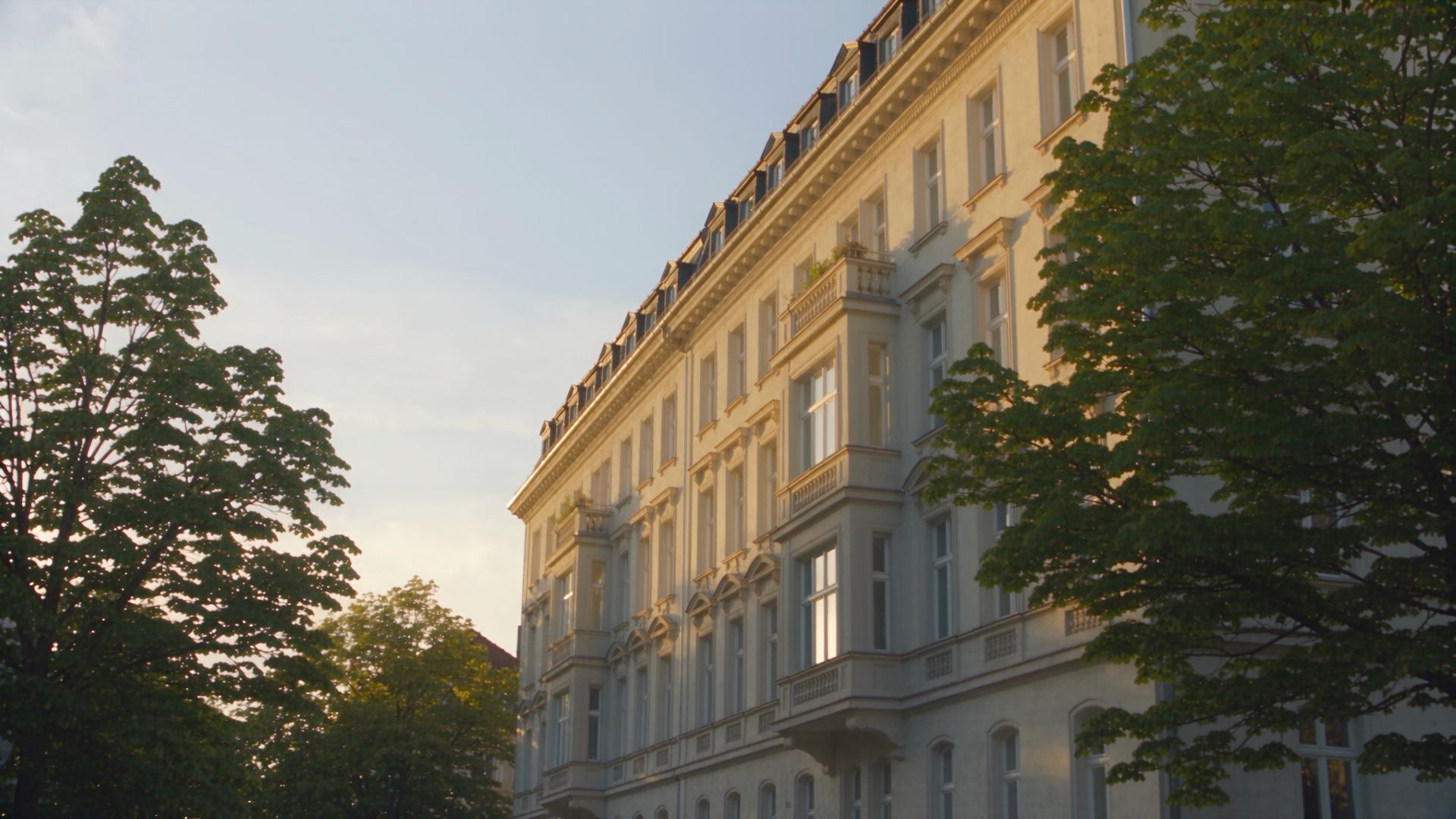 Gründerzeitbau mit heller Stuckfassade und Erkern im warmen Abendlicht, von Bäumen eingerahmt